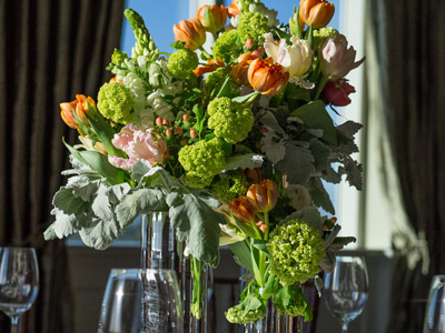 A green, white, and orange floral arrangement with glassware in front of windows.