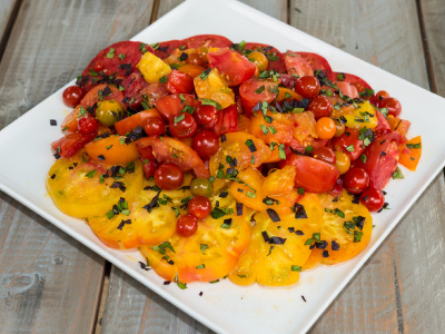 A plate of cut up red, orange, and yellow tomatoes with herbs on a wooden table.