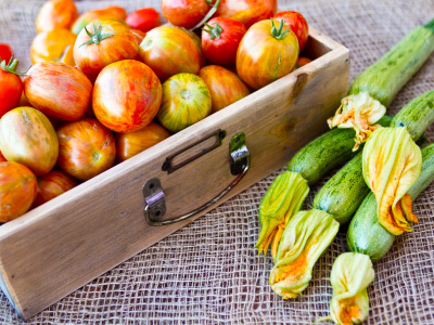 A box filled with red tomatoes next to green zucchini on a burlap bag.