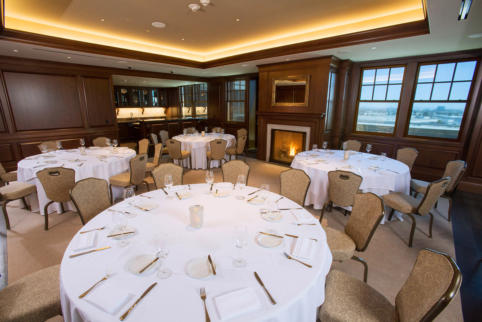 A wood-paneled room with round tables, tan seats, a fireplace, windows, and a kitchen