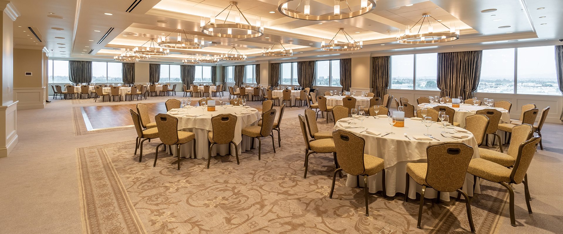 A room in cream, white, and beige with dining tables, a dance floor, chandeliers, and windows