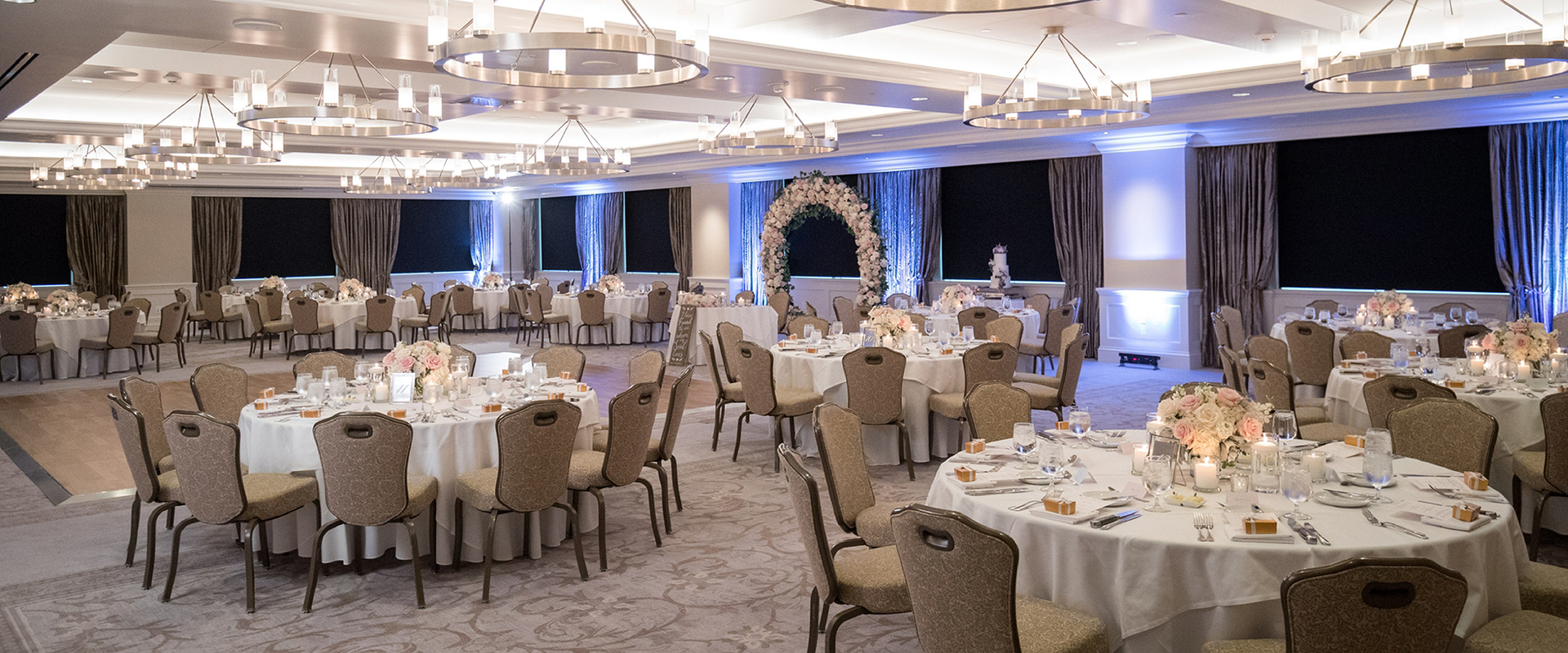 A room in cream, beige, and white with round tables, a dance floor, chandeliers, and windows
