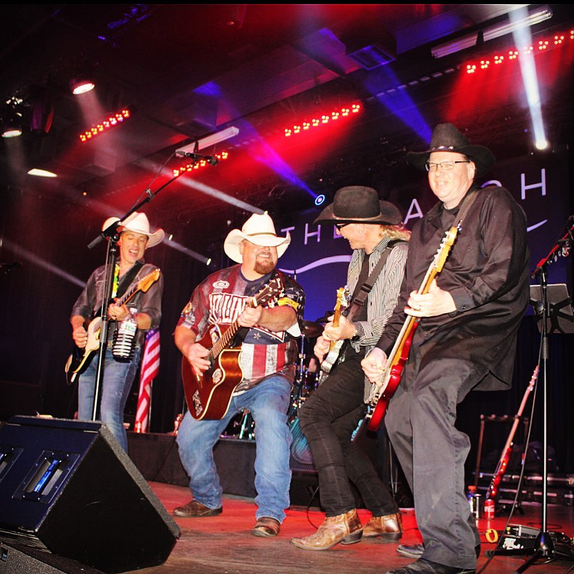 A group of 4 band members playing instruments on a brightly lit stage.