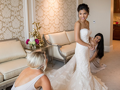 A person wearing a wedding gown stands in a softly lit dressing room while attendants kneel to adjust the lace train, surrounded by elegant seating and a crystal chandelier.