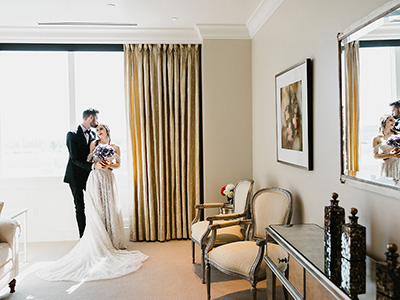 Two people stand near a window holding a bouquet in a softly lit suite, reflected in a wall mirror beside classic chairs and decorative accents.