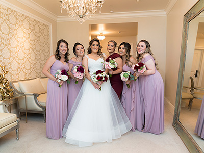 A person wearing a wedding gown stands surrounded by attendants in matching dresses, all holding coordinated bouquets in an elegant dressing room.