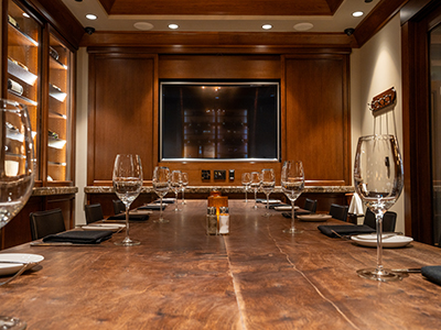 Close-up view down a private dining table set with wine glasses, plates, candles, and a mounted flat screen on a wood-paneled wall