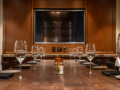 Front-facing view of a private dining table with place settings, wine glasses, candles, and a wall-mounted screen in a wood-paneled room