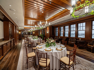 A richly detailed dining room with dark wood paneling, round tables set with white linens and floral centerpieces, wooden chairs, and warm chandelier lighting.