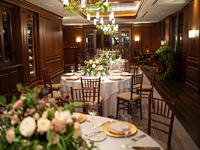 A warmly lit private event space featuring wood-paneled walls, round tables with elegant place settings, lush greenery centerpieces, and classic chandelier fixtures.