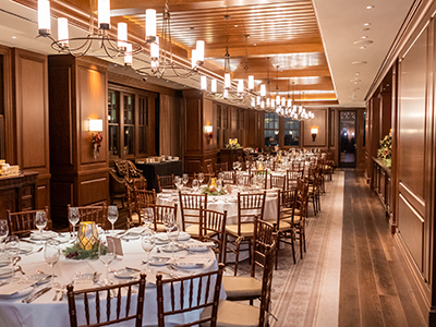 A formal dining setup with round tables, white linens, candlelit centerpieces, wooden chairs, and soft lighting along a wood-paneled interior.
