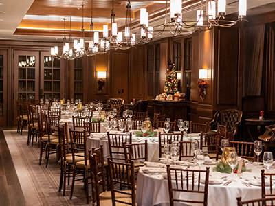 A festive banquet room with round tables, white linens, wooden chairs, holiday décor, and soft ambient lighting beneath modern chandeliers.