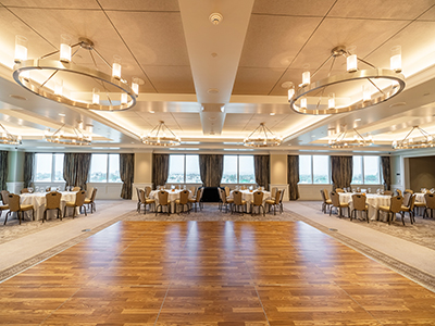 A wide view of a formal event space featuring a polished wood dance floor centered between symmetrically arranged dining tables and contemporary ceiling lights.