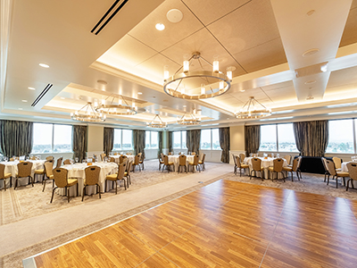 A spacious banquet hall with a wood dance floor, round dinner tables, patterned carpet, and large windows providing natural light.