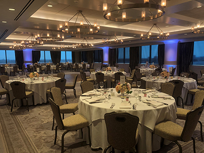 An evening-lit banquet hall with round tables, white linens, floral centerpieces, and subtle blue accent lighting creating a warm event atmosphere.