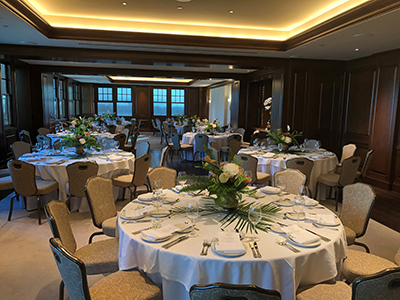 An intimate banquet room with dark wood paneling, round tables dressed in white linens, floral centerpieces, and warm recessed ceiling lighting.