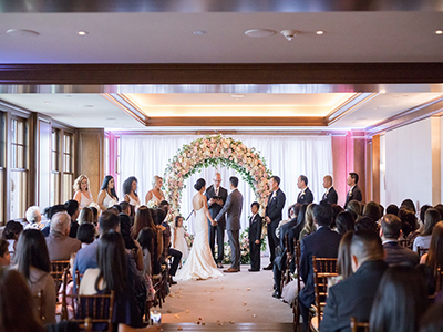 A wedding ceremony in progress with guests seated on either side of an aisle, the couple standing beneath a circular floral arch, and soft pink uplighting against wood-paneled walls.