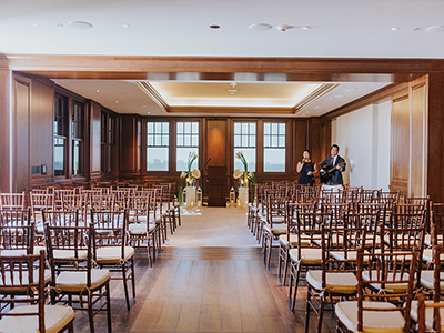 A ceremony setup with wooden chairs arranged in rows facing a minimalist altar framed by tall floral arrangements and lanterns against wood-paneled walls.