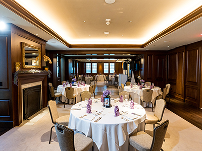 A reception dining room set with round tables, white linens, upholstered chairs, purple table accents, and warm recessed lighting throughout the space.