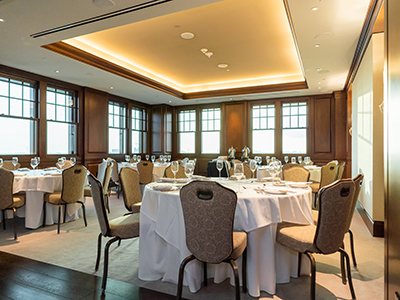 A bright banquet room arranged for dining with round tables, white linens, tall windows, wood paneling, and warm overhead lighting creating an inviting atmosphere.