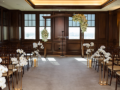 A close view of a ceremony altar featuring white floral arrangements on gold stands, a clear podium, and sunlight streaming through large windows at the front of the room.