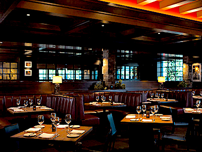 A room with wooden paneling, leather booth seating, a row of wooden tables, lamps, and windows.