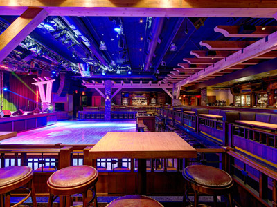 A wooden table with stools overlooking a dance floor, stage and surrounding booth seating