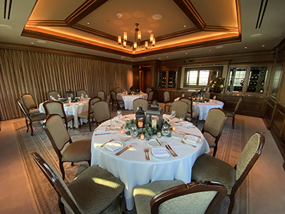 A warmly lit private dining room arranged with multiple round tables dressed in white linens, lantern centerpieces, upholstered chairs, and a coffered ceiling with chandelier lighting.