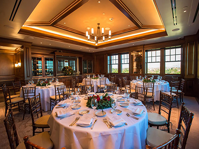 A holiday-themed private dining room with festively set round tables, white linens, wooden chairs, and warm chandelier lighting.