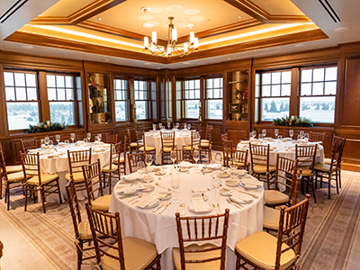 A formal banquet room with round tables, white tablecloths, wooden chairs, seasonal décor, and elegant ceiling chandeliers.